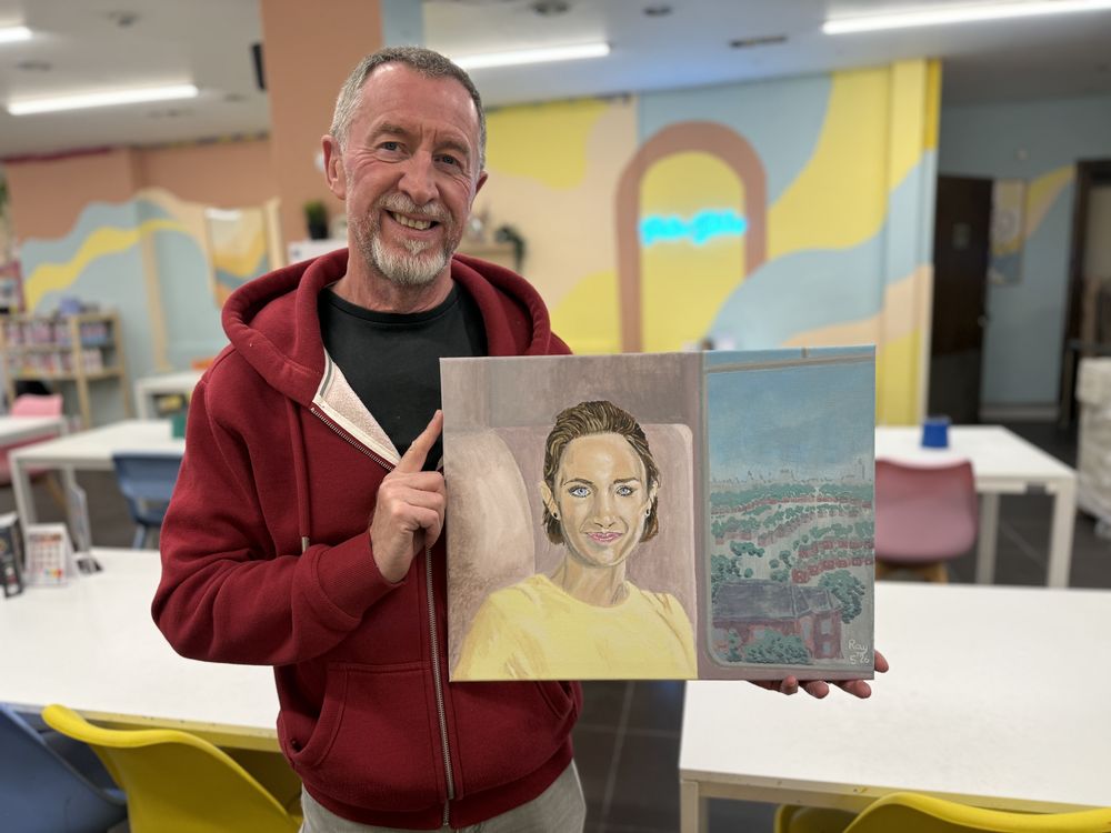 Smiling adult student holding up the portrait and landscape he painted on the course