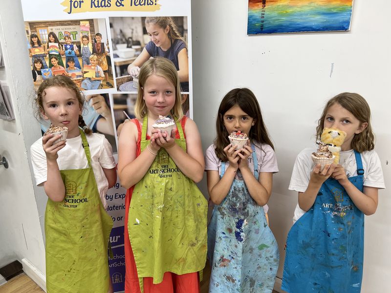 Four children in paint-splashed aprons proudly holding up the clay cupcakes they made in class