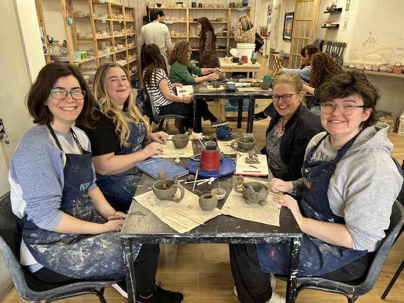 Four smiling adults in aprons around a table of clay at a pottery workshop