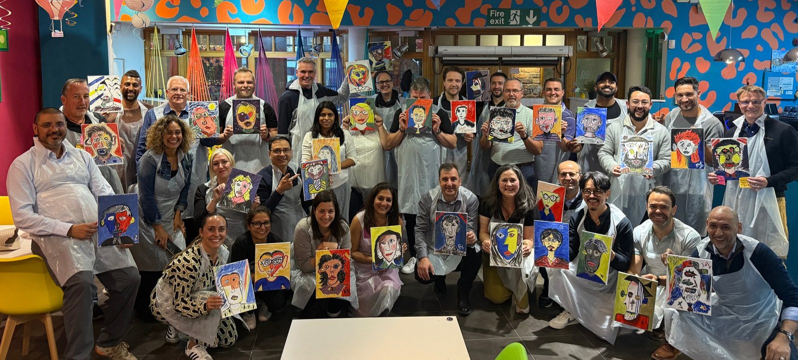 A big corporate group of around 30 colleagues in aprons, each holding up the Picasso-style portrait they painted at their team event