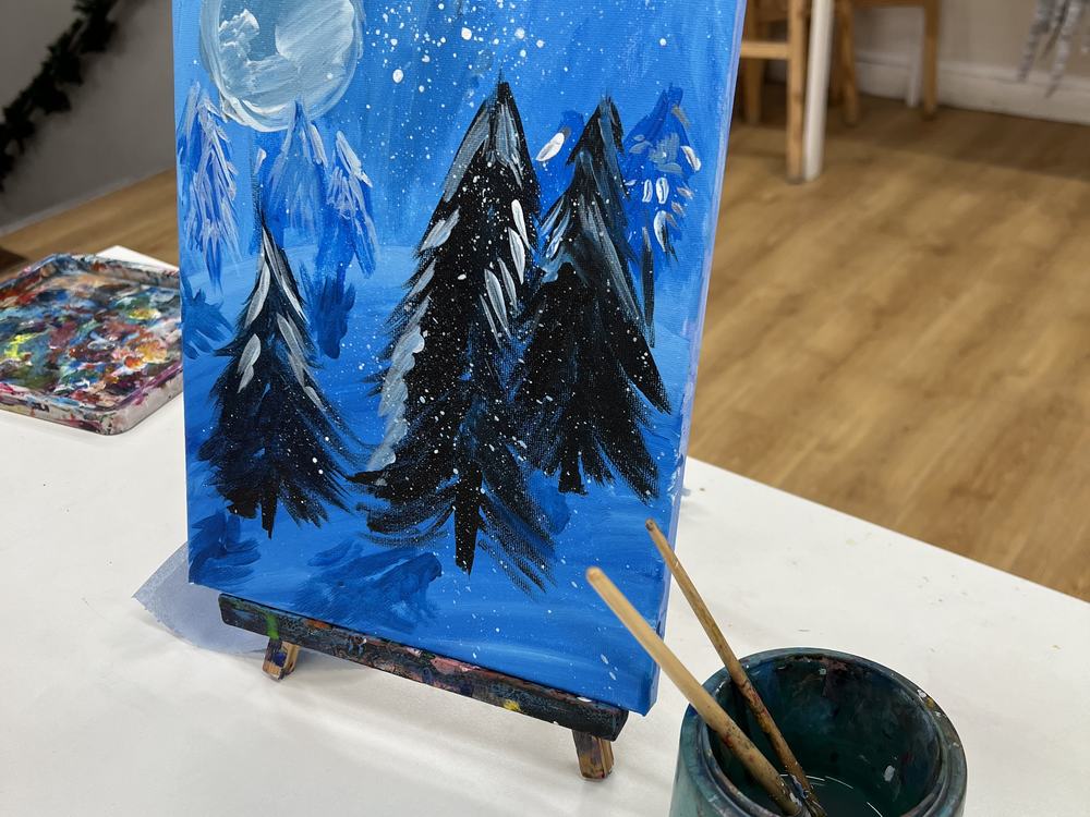 A snowy night-sky canvas with a moon and dark pine trees, painted in the studio, on a small easel