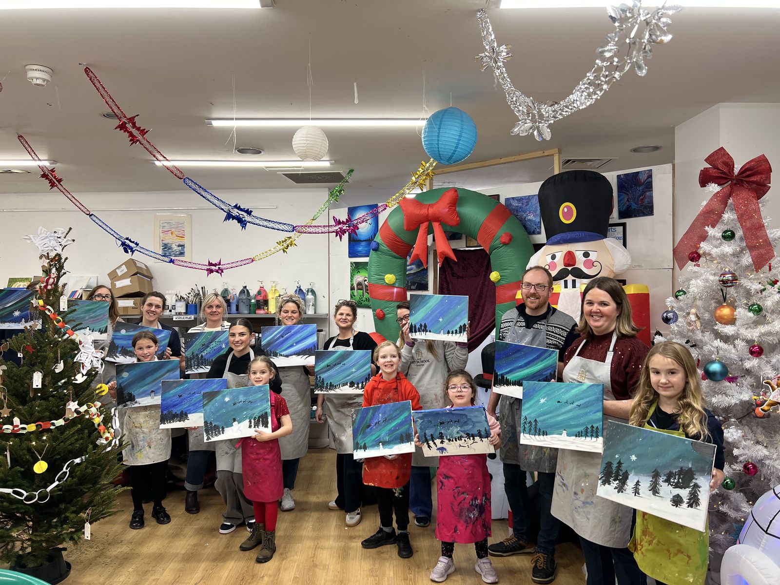 Children and grown-ups holding up their snowy-night canvases in the studio, with Christmas trees and nutcrackers behind them