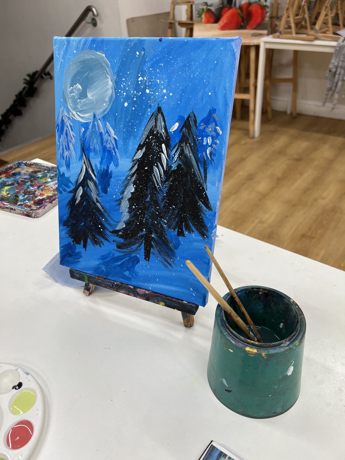 A student’s painted night-sky canvas with a full moon and snowy pine trees on a studio easel